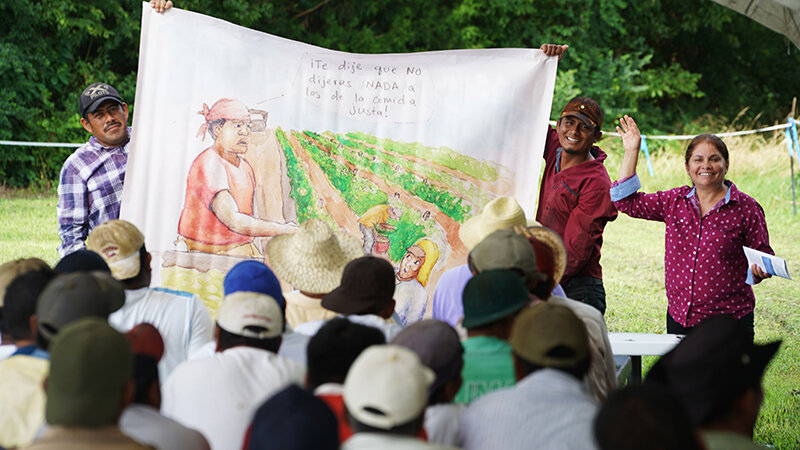 The CIW’s Nely Rodriguez (right) leads a conversation on protection against retaliation under the Fair Food Program during a recent Worker-to-Worker Education session on a Fair Food Program Participating Farm, as two volunteers display an FFP drawing depicting a supervisor scolding a worker for speaking up on the job.